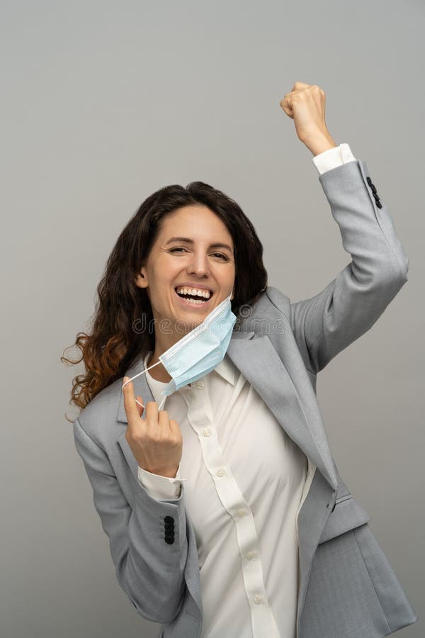 Studio portrait of happy business woman taking off mask, raising fist, grey background. Female removing medical mask from face and looking at camera. Coronavirus, Covid-19 ended. Pandemic is over. Studio portrait of happy business woman taking off mask, raising fist, grey background. Female removing medical mask from face and looking at camera. Coronavirus, Covid-19 ended. Pandemic is over