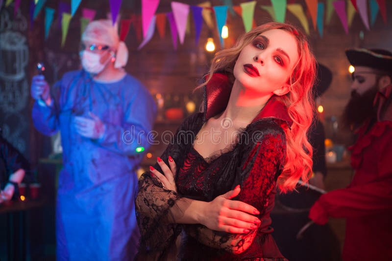 Side view portrait of beautiful young women dressed up like a witch with a serious face at halloween gathering. Man dressed up like a doctor in the background at halloween celebration. Side view portrait of beautiful young women dressed up like a witch with a serious face at halloween gathering. Man dressed up like a doctor in the background at halloween celebration.