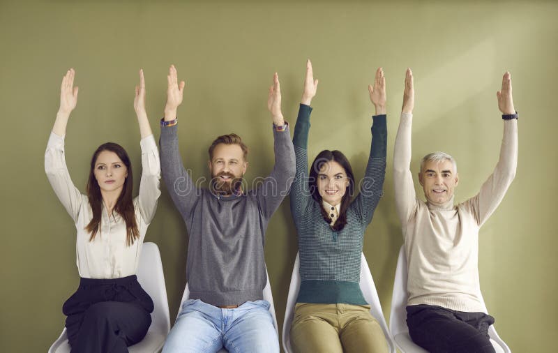 Portrait of people sitting in row on chairs and voting unanimously, raising two hands each. Business people in casual clothes make decisions by voting while sitting against light green wall. Portrait of people sitting in row on chairs and voting unanimously, raising two hands each. Business people in casual clothes make decisions by voting while sitting against light green wall.