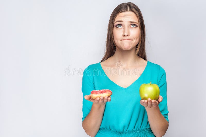 Portrait of dreaming young beautiful girl in blue blouse standing, holding pink donut and green apple in the hands like weighing