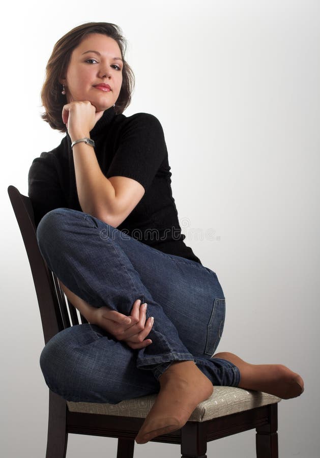Portrait of an attractive young woman posing on a chair; dark hair, dark eyes, dressed in dark blouse and jeans; vertical format; white to gray gradient background; studio;. Portrait of an attractive young woman posing on a chair; dark hair, dark eyes, dressed in dark blouse and jeans; vertical format; white to gray gradient background; studio;