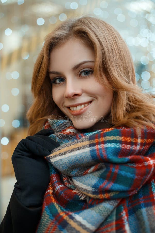 Portrait of a charming woman with blond hair and beautiful smile in a black stylish coat in black gloves with a woolen warm scarf in a cage against the background of garlands. Cute girl. Portrait of a charming woman with blond hair and beautiful smile in a black stylish coat in black gloves with a woolen warm scarf in a cage against the background of garlands. Cute girl