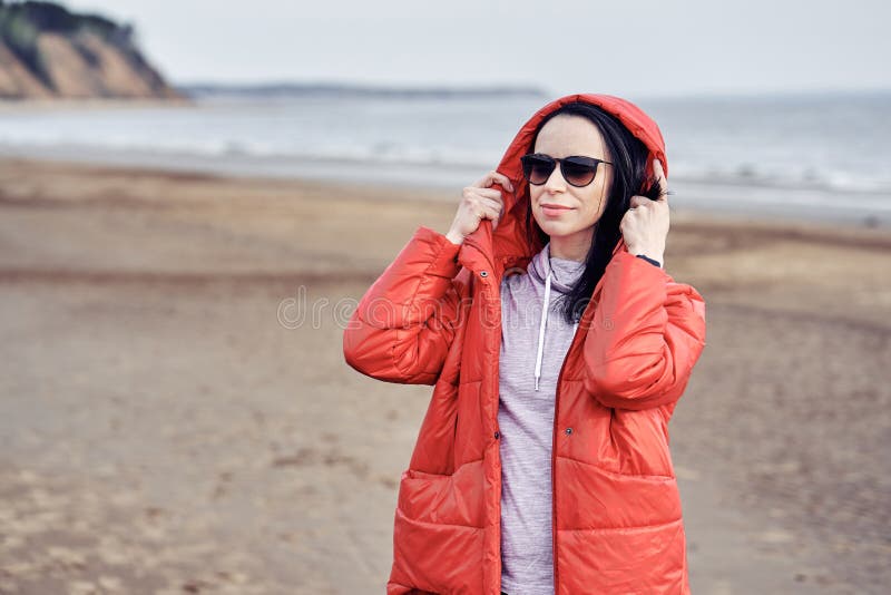 Portrait einer jungen brunette Frau in der Sonnenbrillen und in einer roten Jacke Frau auf einem Weg auf dem sandigen Ufer von ei stockfoto