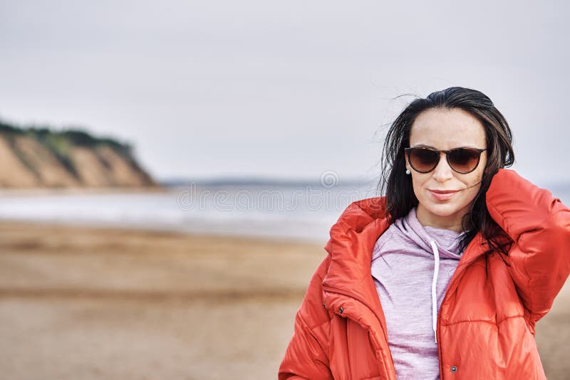 Portrait einer jungen brunette Frau in der Sonnenbrillen und in einer roten Jacke Frau auf einem Weg auf dem sandigen Ufer von ei lizenzfreie stockfotografie