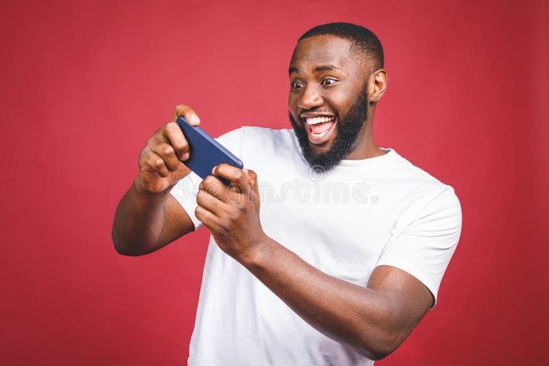 Portrait of a cheerful young handsome  african man dressed in casual playing games on mobile phone isolated over red background. Portrait of a cheerful young handsome  african man dressed in casual playing games on mobile phone isolated over red background