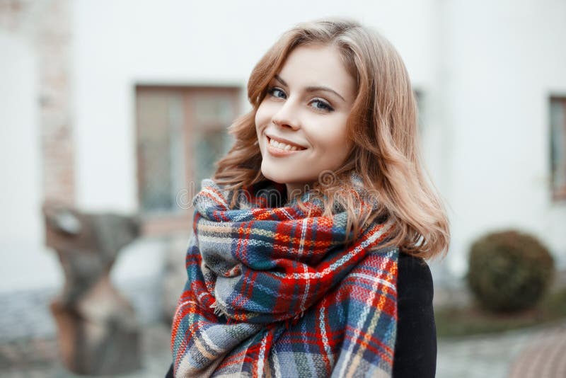 Portrait of a cute young girl with beautiful blue eyes with an attractive smile with curly blond hair in winter stylish clothes on the background of a modern building. Charming girl. Portrait of a cute young girl with beautiful blue eyes with an attractive smile with curly blond hair in winter stylish clothes on the background of a modern building. Charming girl