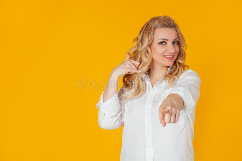 Call me Maybe. Portrait of a caucasian blonde woman smiling pointing to the camera, cheeky smiling and making a phone gesture, as if asking for a number, flirting with someone, yellow background. Call me Maybe. Portrait of a caucasian blonde woman smiling pointing to the camera, cheeky smiling and making a phone gesture, as if asking for a number, flirting with someone, yellow background