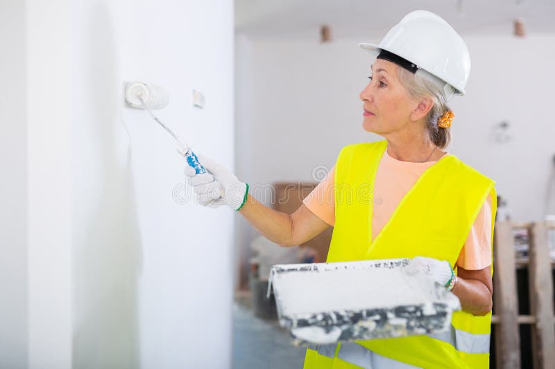 Portrait of elderly positive woman painter in protective helmet and yellow vest who paints the walls with paint roller. Portrait of elderly positive woman painter in protective helmet and yellow vest who paints the walls with paint roller