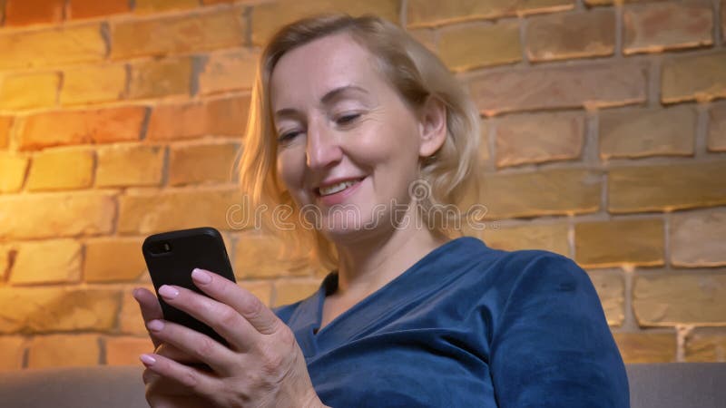 Portrait en gros plan de la dame caucasienne supérieure joyeuse s'asseyant sur le sofa et observant dans le smartphone en atmosph photographie stock libre de droits Portrait en gros plan de la dame caucasienne supérieure joyeuse s'asseyant sur le sofa et observant dans le smartphone en atmosph photographie stock libre de droits