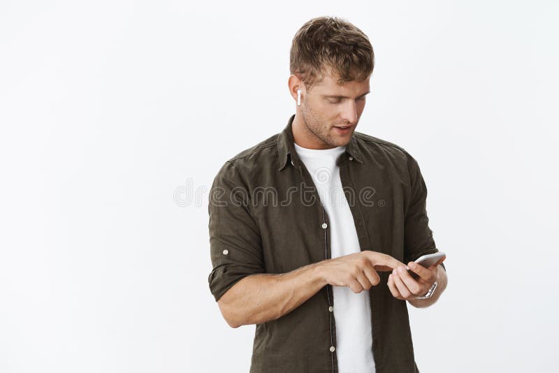 Portrait of focused good-looking guy in wireless white earbuds searching right music to sute mood standing over gray background touching screen and looking at mobile phone, picking song.