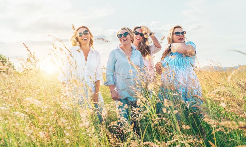 Portrait of four cheerful smiling and laughing women embracing during outdoor walking by high green grass hill. They looking at