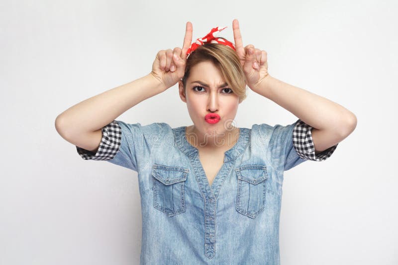 Portrait of funny beautiful young woman in casual blue denim shirt with makeup and red headband standing with horns hands on head