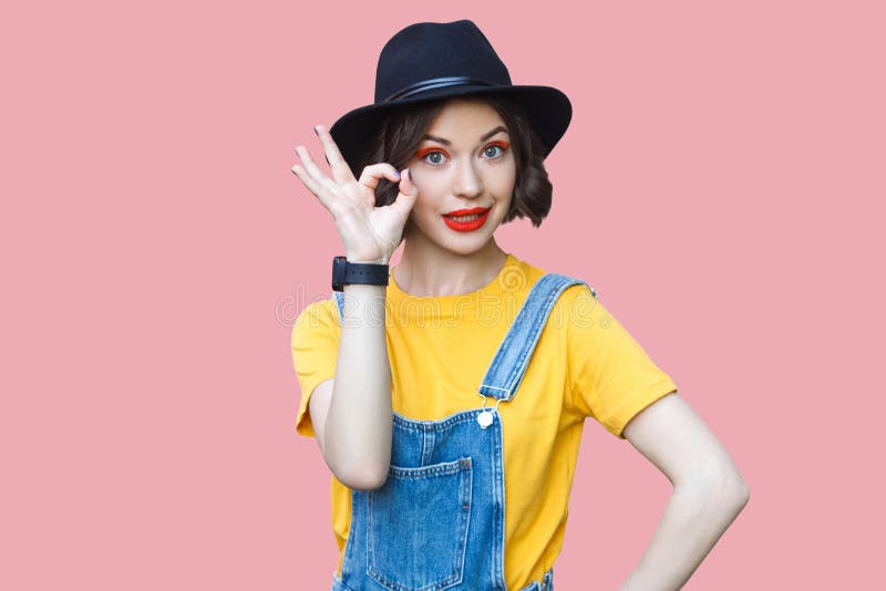 Portrait of funny beautiful young woman in yellow t-shirt and blue denim overalls with makeup and black hat standing with ok sign stock photos Portrait of funny beautiful young woman in yellow t-shirt and blue denim overalls with makeup and black hat standing with ok sign stock photos