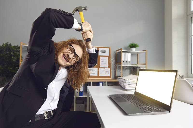 Portrait of a funny crazy office worker sitting at a desk breaking a laptop with a hammer. stock image