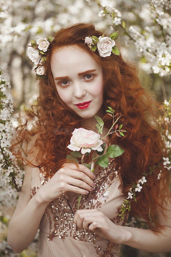 Portrait of Gentle woman with long red hair with a rose in a blooming garden. Red-haired girl with pale skin and blue