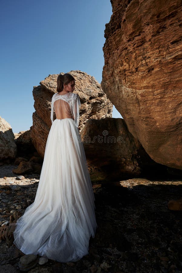 Portrait of a girl in a wedding gorgeous dress posing a photographer on the beach. The bride is on the rocks stock photo Portrait of a girl in a wedding gorgeous dress posing a photographer on the beach. The bride is on the rocks stock photo