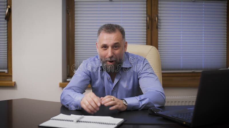 Portrait of a good-natured businessman. Director in a blue shirt sitting at office desk, smiling and looking at the