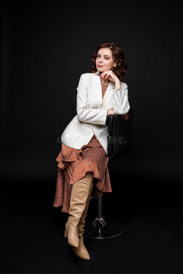 Portrait of handsome woman in long dress and boots sits on a chait and poses for the camera, picture isolated on black