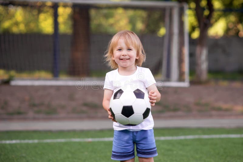 Portrait of happily smiling blonde toddler boy standing at stadium holding ball against soccer nets stock photo Portrait of happily smiling blonde toddler boy standing at stadium holding ball against soccer nets stock photo