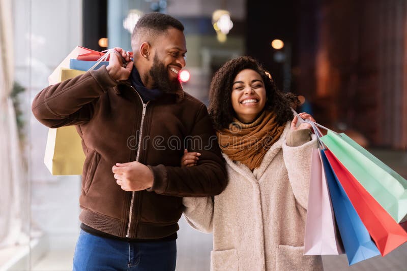 Love And Relationship Concept. Portrait of cheerful african american woman laughing, black couple walking down the street in the evening, holding shopping bags, having good time and fun. Love And Relationship Concept. Portrait of cheerful african american woman laughing, black couple walking down the street in the evening, holding shopping bags, having good time and fun
