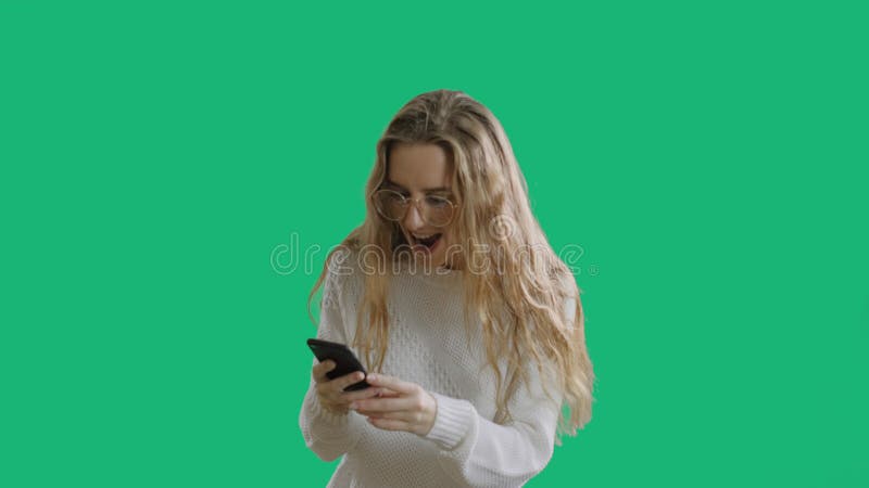 Portrait of a happy beautiful girl in glasses is looking to a mobile phone and celebrating at green screen chroma key