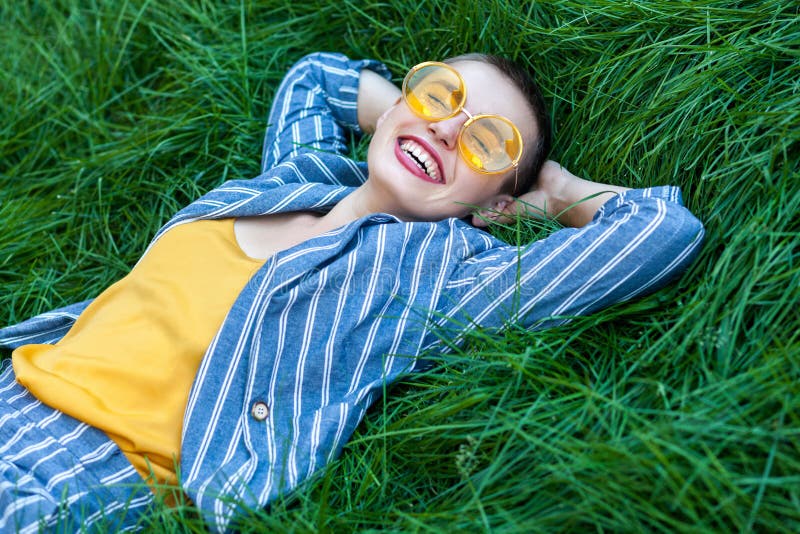 Portrait of Happy fancy young woman with short hair in casual blue striped suit, yellow shirt, glasses lying down on green grass