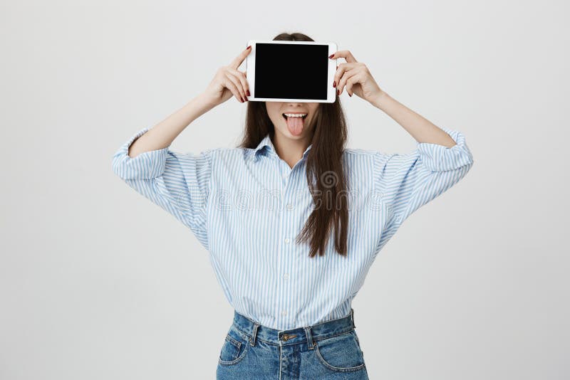 Portrait of happy and funny young european student covering face with tablet and sticking out tongue, standing over gray stock images Portrait of happy and funny young european student covering face with tablet and sticking out tongue, standing over gray stock images