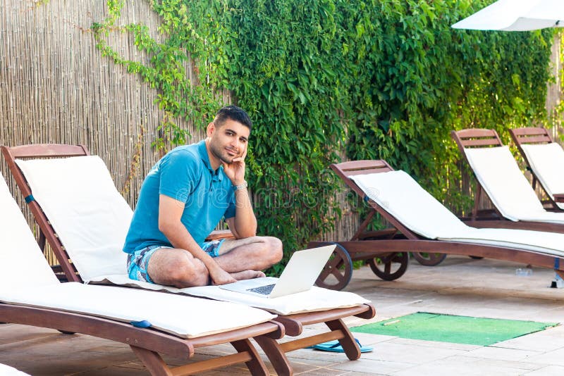 Portrait of happy handsome bearded young adult freelancer man in blue t-shirt and shorts sitting on deckchair with laptop on royalty free stock photography Portrait of happy handsome bearded young adult freelancer man in blue t-shirt and shorts sitting on deckchair with laptop on royalty free stock photography