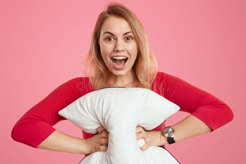 Portrait of happy light haired young woman with overjoyed facial expression, holds soft white pillow, dressed in red jumper, opens stock photo Portrait of happy light haired young woman with overjoyed facial expression, holds soft white pillow, dressed in red jumper, opens stock photo