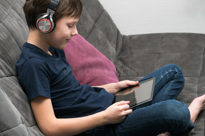 Happy boy with tablet computer resting on sofa, side view. Child in headphones play videogame on gadget or watch video.