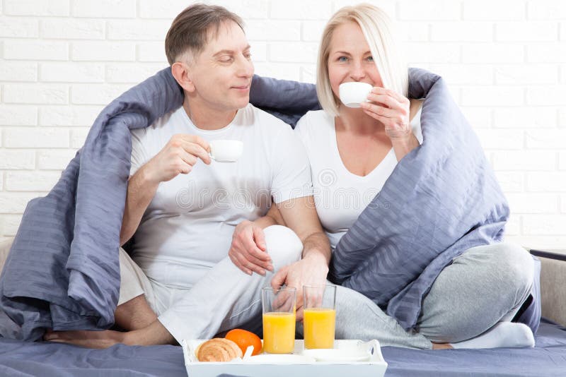 Portrait of happy playful couple relaxing in comfortable cozy bed looking at each other, smiling man and woman having fun posing. Holding cups of coffee while covering their shoulders with a blanket. Portrait of happy playful couple relaxing in comfortable cozy bed looking at each other, smiling man and woman having fun posing. Holding cups of coffee while covering their shoulders with a blanket.