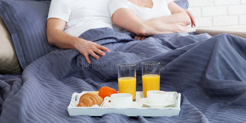 Portrait of happy playful couple relaxing in comfortable cozy bed looking at each other, smiling man and woman having fun posing. Holding cups of coffee while covering their shoulders with a blanket. Portrait of happy playful couple relaxing in comfortable cozy bed looking at each other, smiling man and woman having fun posing. Holding cups of coffee while covering their shoulders with a blanket.