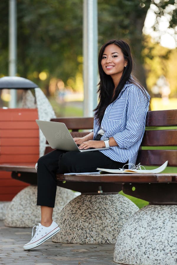 Portrait of a happy pretty asian female student holding laptop computer while sitting on the bench at the park