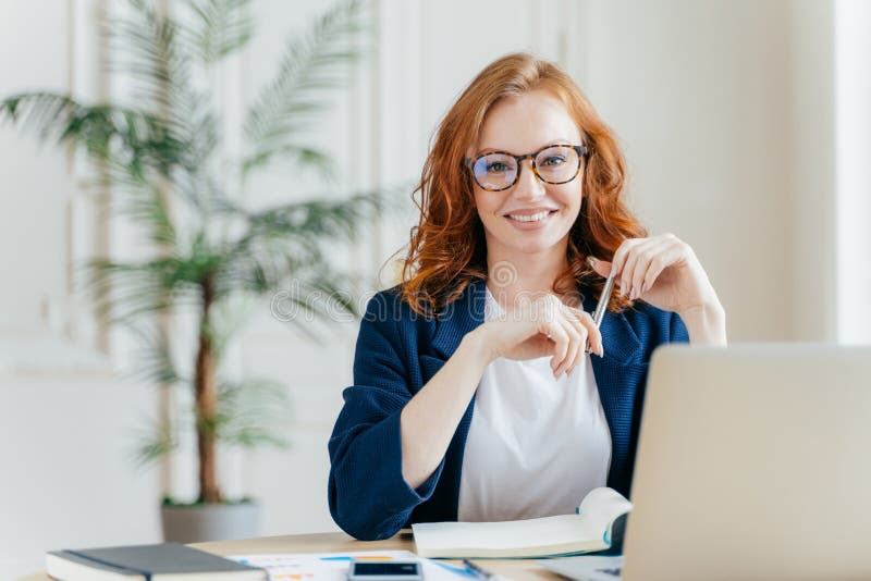 Portrait of happy redhaired woman employee in optical glasses, has satisfied expression, works with modern gadgets, waits for