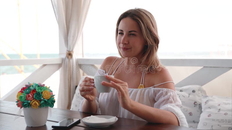 Portrait of happy young beautiful woman drinking coffee in restaurant on the seashore.