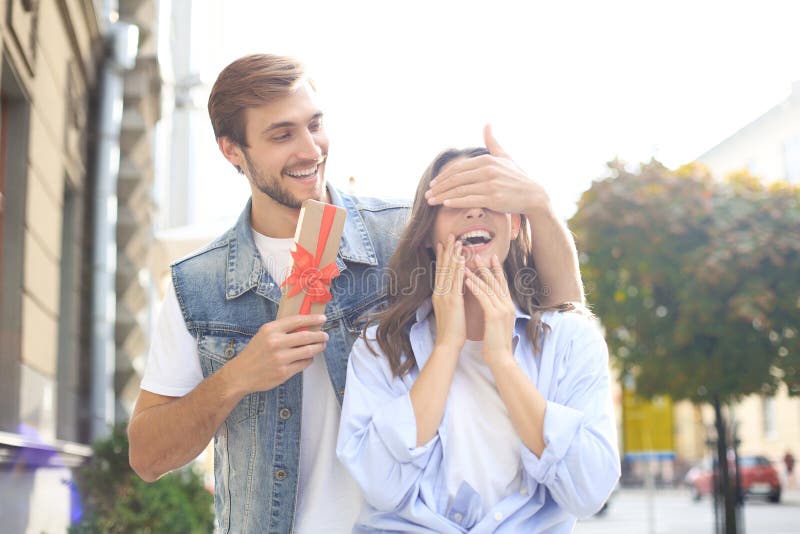Portrait of a happy young couple hugging while standing together with a present box, man covers woman`s eyes. stock photography Portrait of a happy young couple hugging while standing together with a present box, man covers woman`s eyes. stock photography
