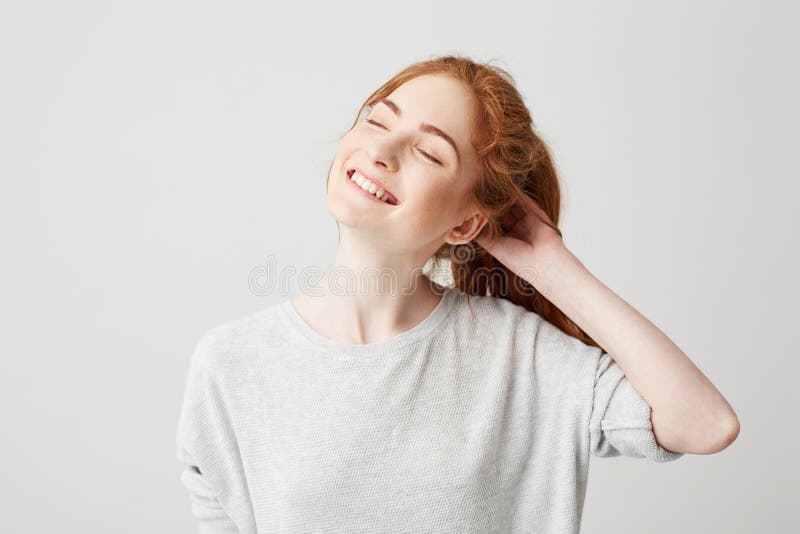 Portrait of happy young cute redhead girl smiling with closed eyes touching hair over white background.