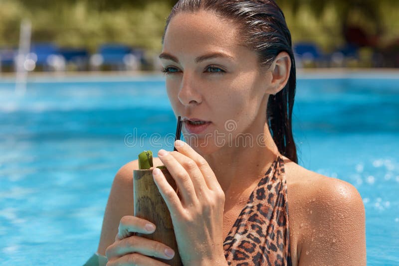 Portrait haut étroit de la position modèle de brune magnétique réfléchie dans la piscine, tenant le conteneur avec le cocktail d' photos stock Portrait haut étroit de la position modèle de brune magnétique réfléchie dans la piscine, tenant le conteneur avec le cocktail d' photos stock