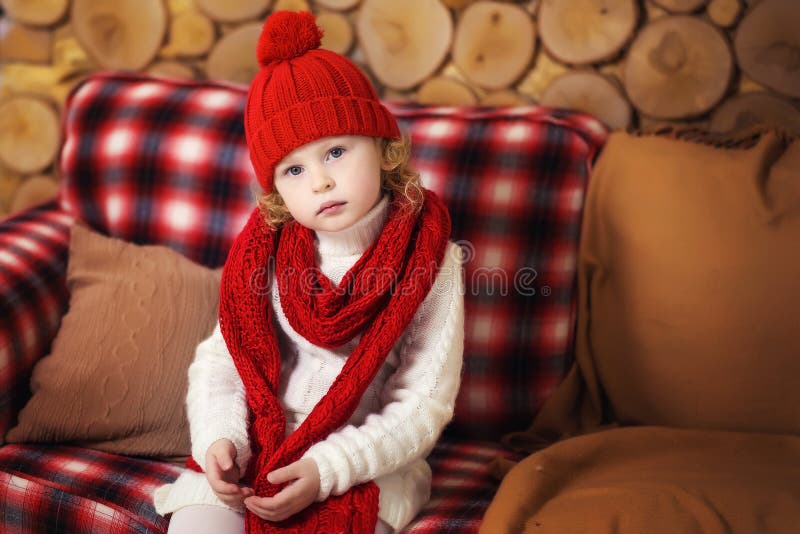 Portrait of little beautiful 3 years old curly blonde girl sitting on sofa in red hat and scarf in wooden room. Winter portrait of