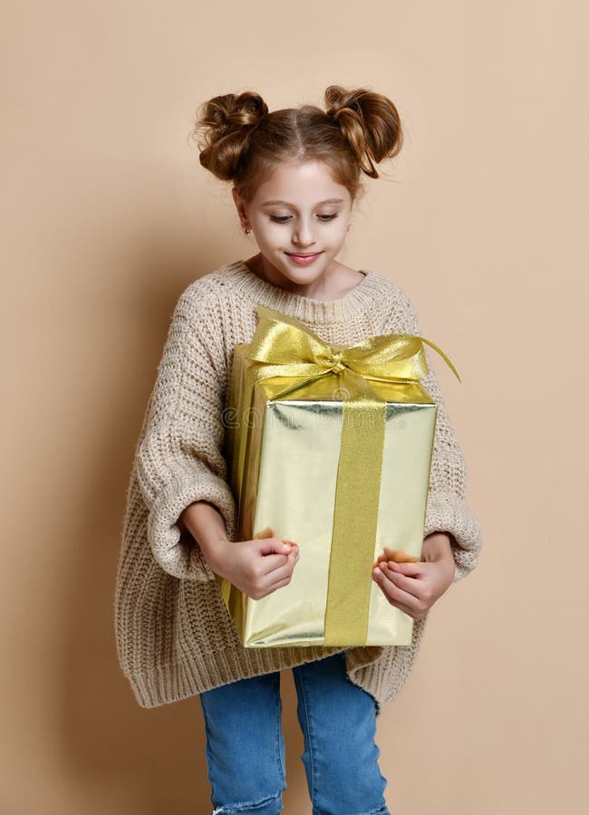 Happy child girl with gift box. Portrait of a little girl in a warm nude knitted sweater on a beige background. A child holds a gold box with a gift at arms. The stock photo Happy child girl with gift box. Portrait of a little girl in a warm nude knitted sweater on a beige background. A child holds a gold box with a gift at arms. The stock photo