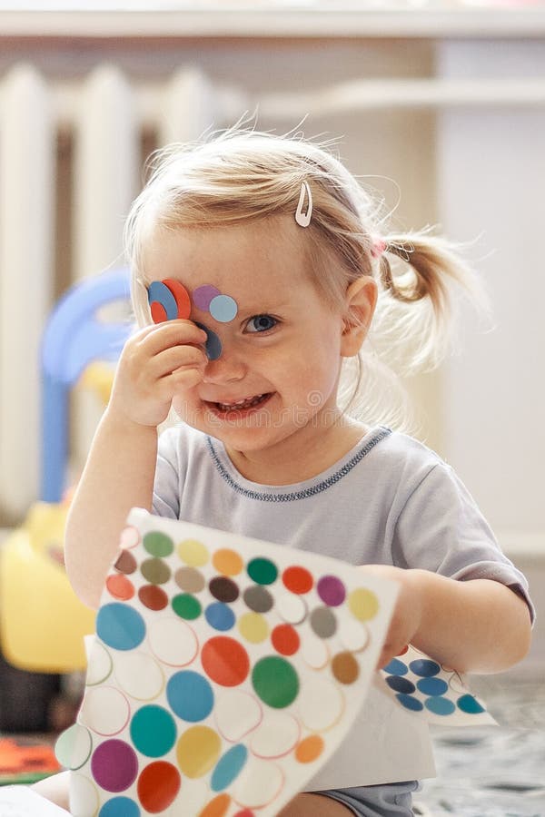 Portrait of a little lovely girl. Cute toddler sticks stickers on herself. Lovely blonde baby in a blue t-shirt.