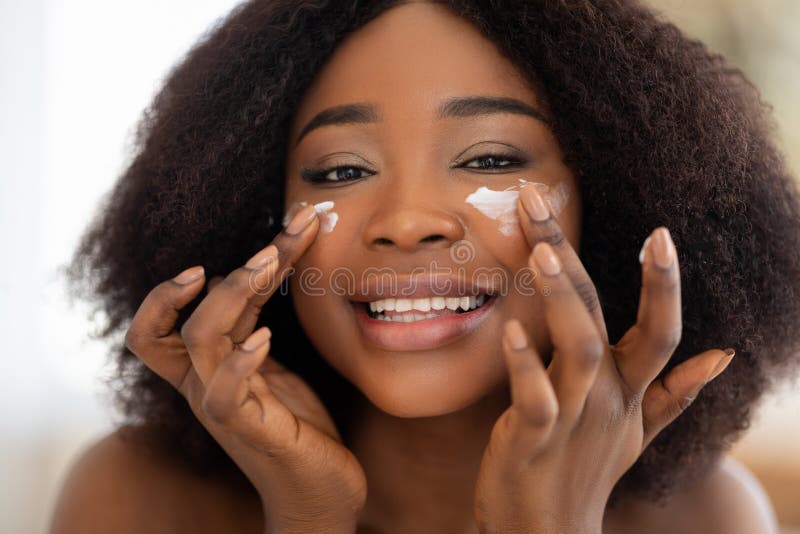 Portrait of lovely black woman applying face cream onto her cheeks, smiling at camera, indoors