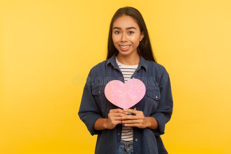 Portrait of lovely girl in denim shirt showing pink heart romantic symbol and looking at camera with toothy smile