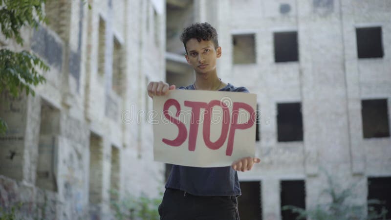 Portrait of male emigrant or refugee posing with Stop banner at the background of neglected urban building. Young