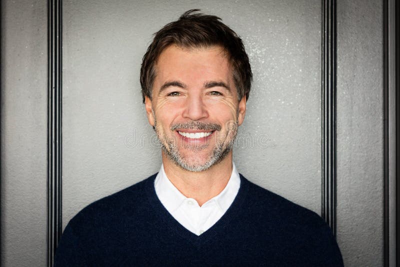 A portrait of a middle-aged bearded man smiling at the camera on a gray background