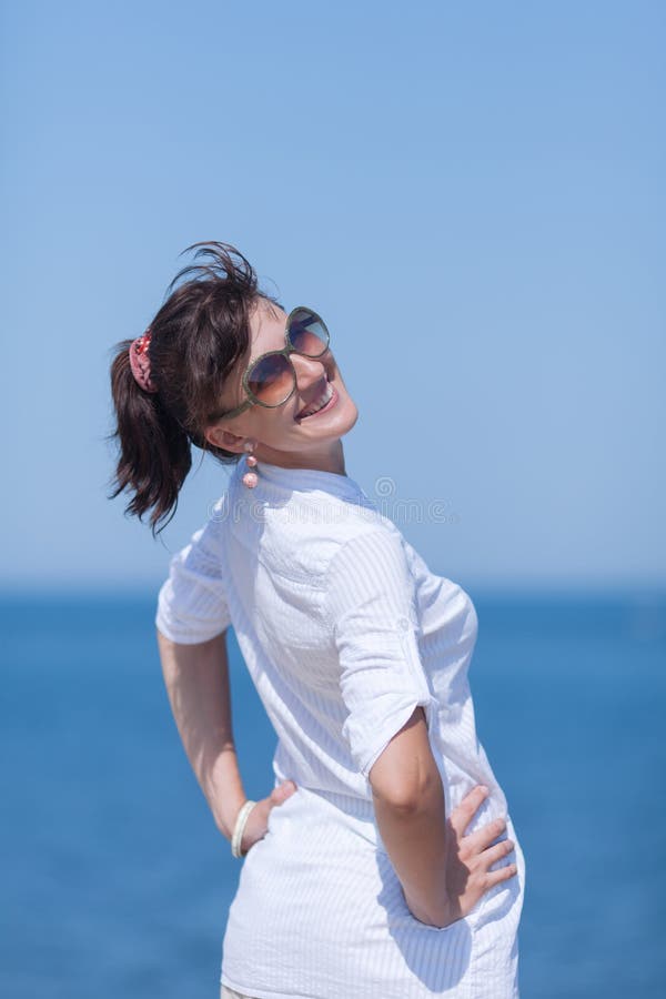 Portrait of middle aged woman against sea. Girl in white stands with arms akimbo and looks over shoulder smiling