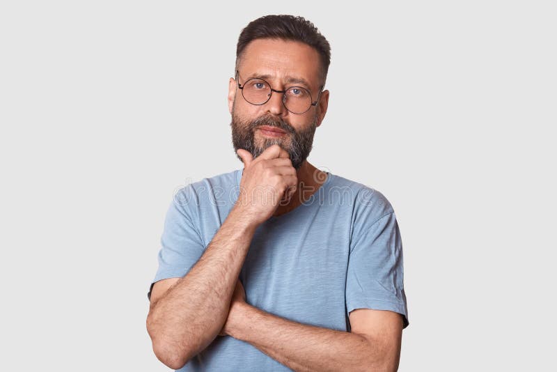 Portrait of middle bearded aged male with pensive facial expression, dressed gray cassual t shirt and round spectacles, keeps hand royalty free stock photo Portrait of middle bearded aged male with pensive facial expression, dressed gray cassual t shirt and round spectacles, keeps hand royalty free stock photo