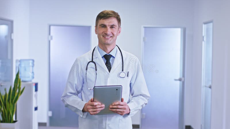 Portrait moderne de couloir d'hôpital d'un homme de médecin avec une tablette numérique donnant directement à la caméra et au sour