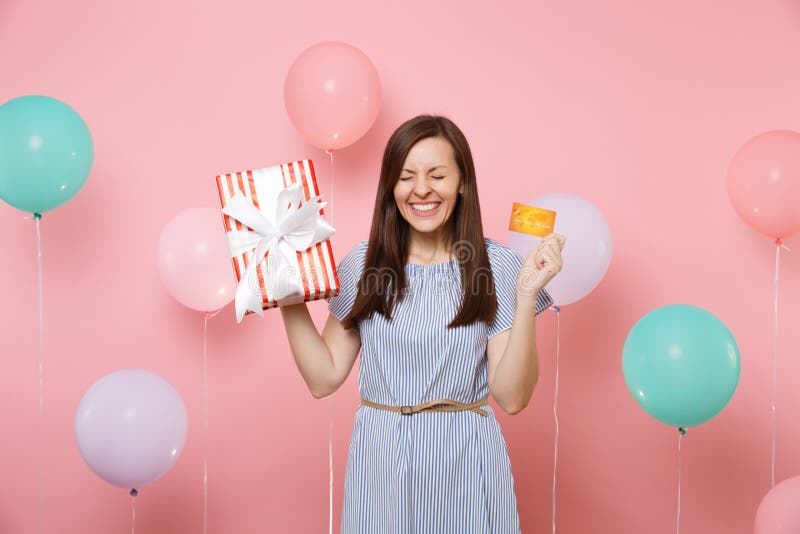 Portrait of overjoyed woman with closed eyes in blue dress hold credit card and red box with gift present on pink