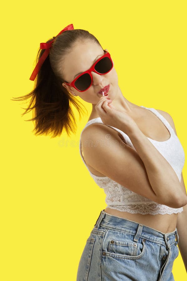 Portrait of playful young woman in sunglasses eating lollipop over yellow background